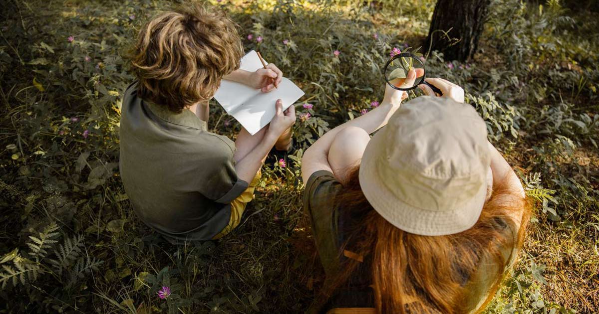 Two students doing a nature study at a summer camp, one sketching in a notebook while the other examines a wildflower with a magnifying glass